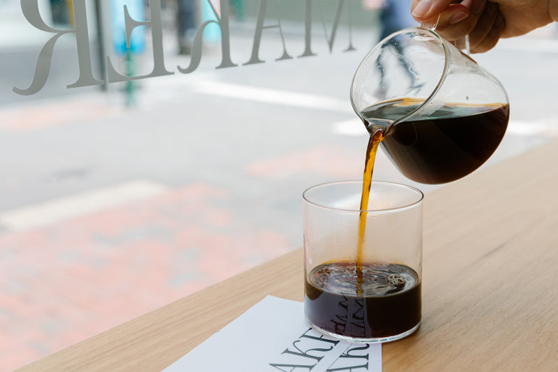 Coffee being poured from a glass carafe into a clear glass on a wooden table.
